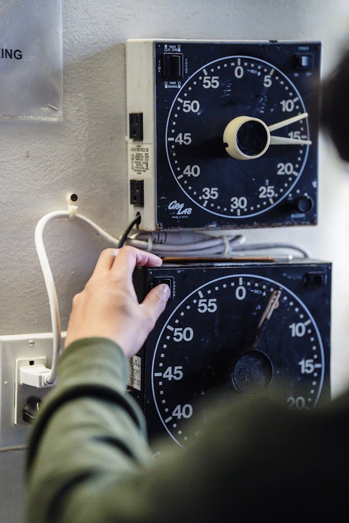Photo by Annushka Ahuja A hand adjusts vintage analog timer gauges mounted on a wall. Vertical shot.